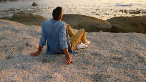 Rückansicht-Eines-Gemischtrassigen-Mannes,-Der-Bei-Sonnenuntergang-Auf-Einem-Felsen-Am-Strand-Sitzt,-4k