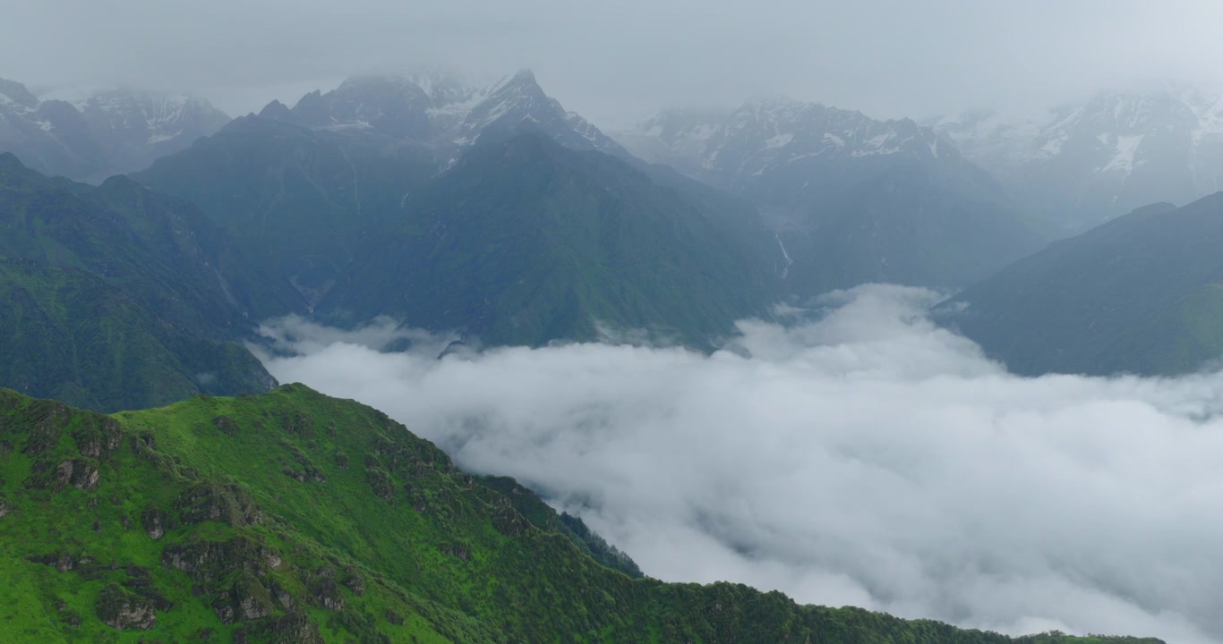 Panchpokhari, Sindhupalchowk, Nepal, tomada ampla de drone deslizando sobre cumes de montanhas verdes com uma densa inversão de nuvens brancas preenchendo o vale abaixo e picos irregulares do Himalaia salpicados de neve.
