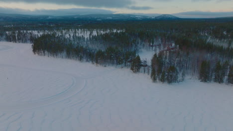 Schneebedeckte-Spuren-Auf-Norbotten-Ice-Lake-Course-Mit-Lappland-Waldland-Und-Bergkette-Sonnenaufgang-Skyline-Aus-Der-Luft