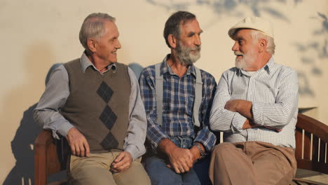 Portrait-Of-The-Three-Old-Men-Sitting-On-A-Bench-And-Posing-To-The-Camera-With-Smiles-At-The-Wall