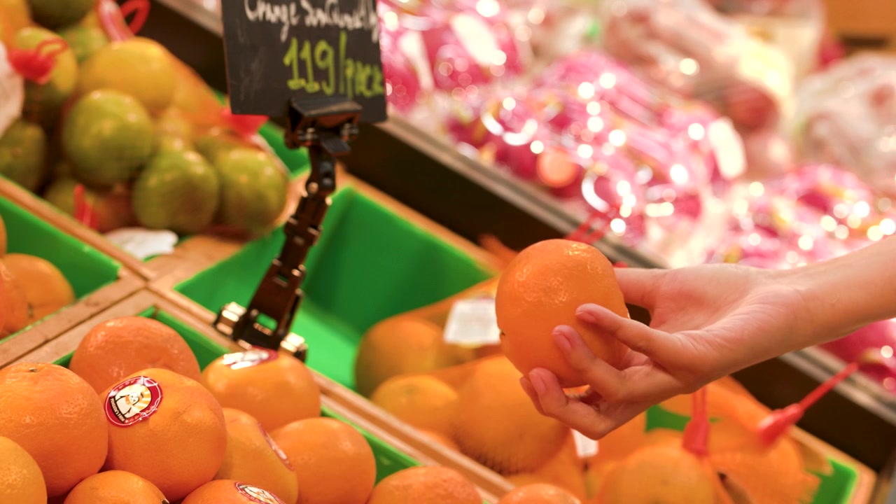 Close-up shot of a hand picking up and inspecting an orange in a supermarket.