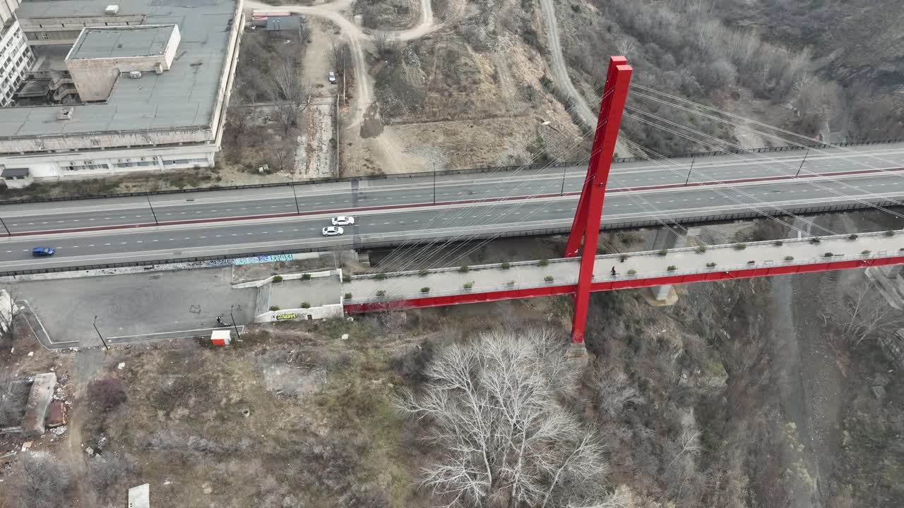 A top-down aerial view of a bold red bridge spanning over a road and surrounded by sparse winter trees and urban outskirts. The structural design stands out in its urban-rural blend