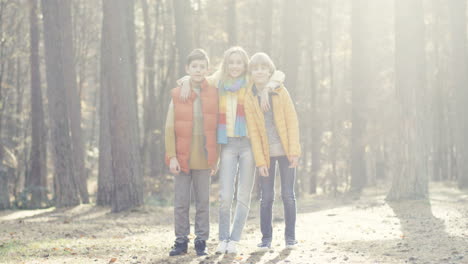 distant view of joyful kids, girl and two boys, hugging and smiling at camera in the forest on a sunny day