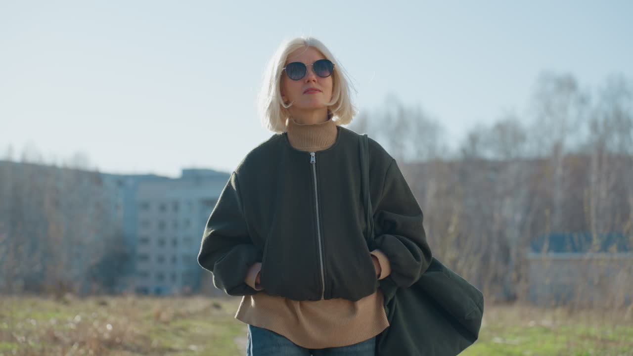 Confident blonde woman in sunglasses wearing dark jacket and turtleneck walking outdoors in sunny urban environment with buildings and trees in background, enjoying lifestyle moment in natural light