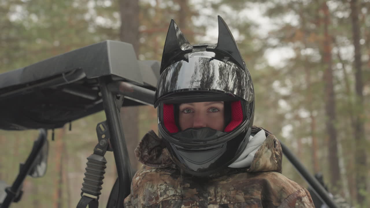 Primo piano di un pilota caucasico con casco, giacca mimetica e occhiali rossi, seduto accanto a un ATV blu in pineta, guanti invernali, sguardo concentrato, test dell'equipaggiamento e preparazione per l'off-road, cielo coperto.