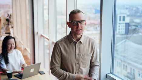 mid-aged man wearing glasses stands at window. man turns to a female colleague and gestures laughing.