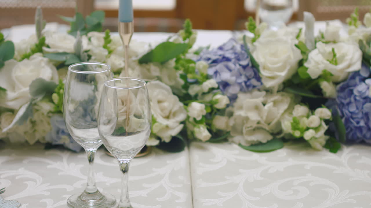 A detailed clip of an elegantly decorated wedding table. It features stemware, a white tablecloth, plates, cutlery, cloth napkins, a delicate floral arrangement, and a personalized invitation.