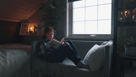 girl picking up a book and makes herself comfortable on a couch and starts reading. slight camera pan