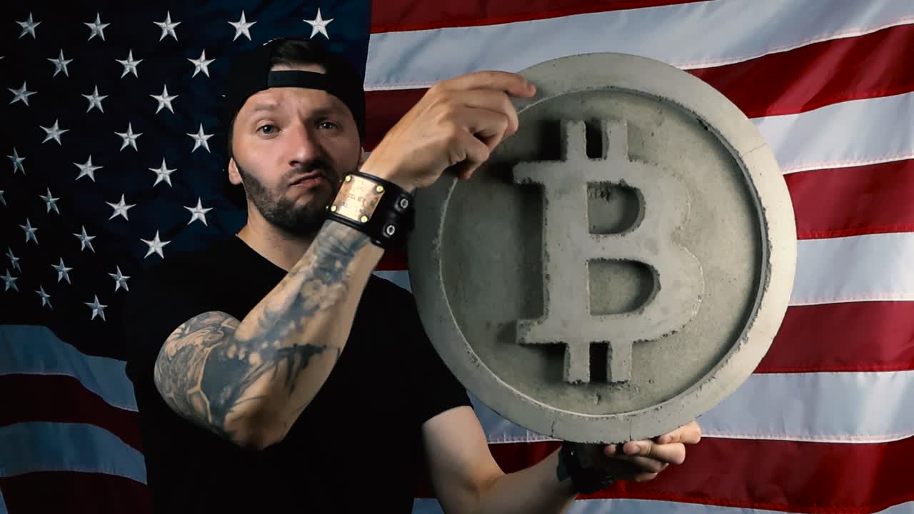 Man with tattooes, beard and black clothing lifting up a large gray bitcoin concrete sculpture and begins to shake his head in front of an American US flag showing he is not optimistic and happy.