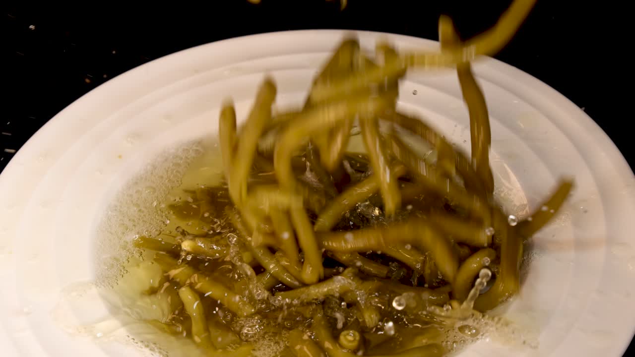 Cooked green beans are dropped into a white bowl filled with broth, captured in a top-down view with bright, even lighting and minimal camera movement
