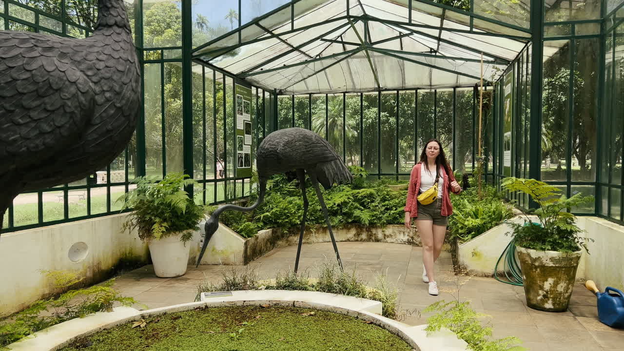 A young woman, wearing a bright yellow fanny pack, walks through the classic glass and metal greenhouse (Estufa de Samambaias) with two distinctive black crane statues and lush ferns