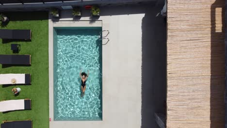 drone flies over an outdoor pool in bird's eye view - woman in swimming costume floats on her back on water - villa in greece crete with palm trees