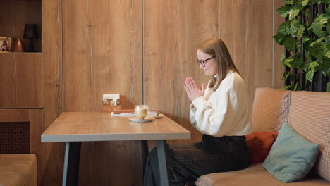 happy woman seated at wooden cafe table smiling and clapping hands in front of latte and chocolate cake, expressing excitement and gratitude before enjoying food in cozy modern interior