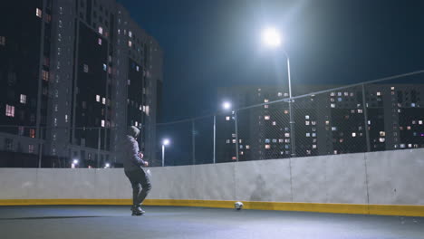 vista trasera de un hombre con capucha jugando al fútbol contra la pared bajo la iluminación brillante del estadio por la noche, la pelota de fútbol cerca de la pared refleja la silueta con el paisaje urbano y los edificios de apartamentos iluminados