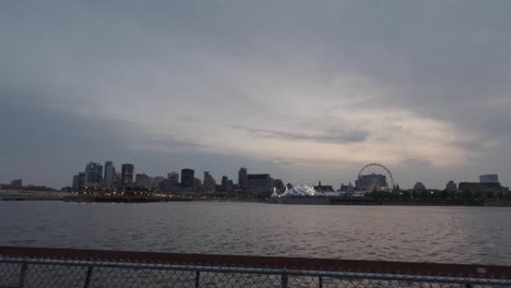 Zeitrafferaufnahme-Der-Skyline-Der-Innenstadt-Von-Montreal-Von-Der-Anderen-Seite-Des-Wassers-Von-Sonnenuntergang-Bis-Nacht,-Mit-Wunderschönem-Himmel,-Lichtern-Der-Stadt-Und-Einem-Zaun-Im-Vordergrund