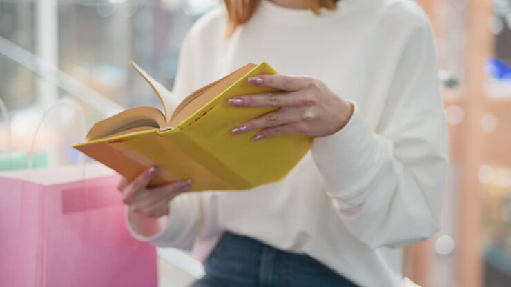 Close-up of young woman flipping through yellow hardcover book while seated near pink shopping bag in modern mall, delicate hands and nails add elegance, with soft background