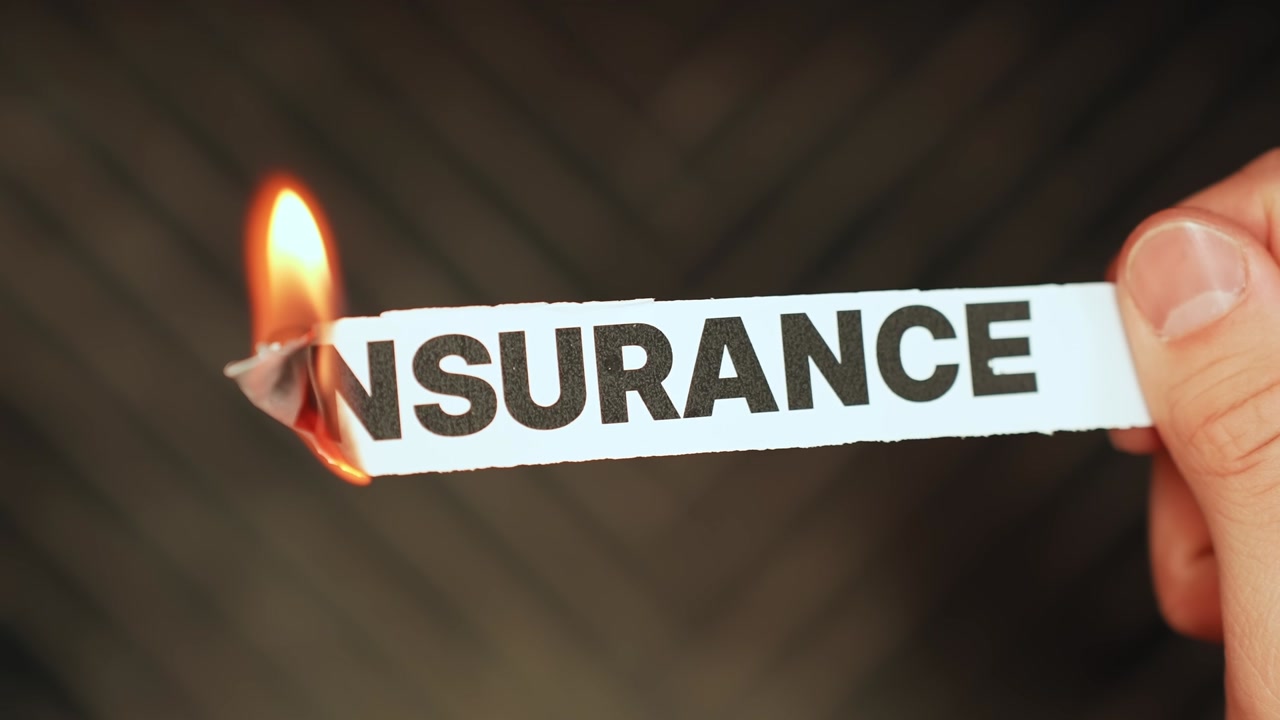 Hand burns a paper label reading INSURANCE, symbolizing policy cancellation, no coverage, uninsured risk, and rising financial uncertainty.
