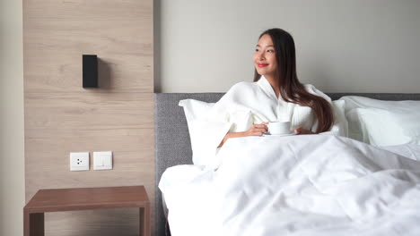 Happy-Attractive-Female-in-Bathrobe-Enjoying-Her-Morning-Coffee-in-Bed-Looking-Through-Window