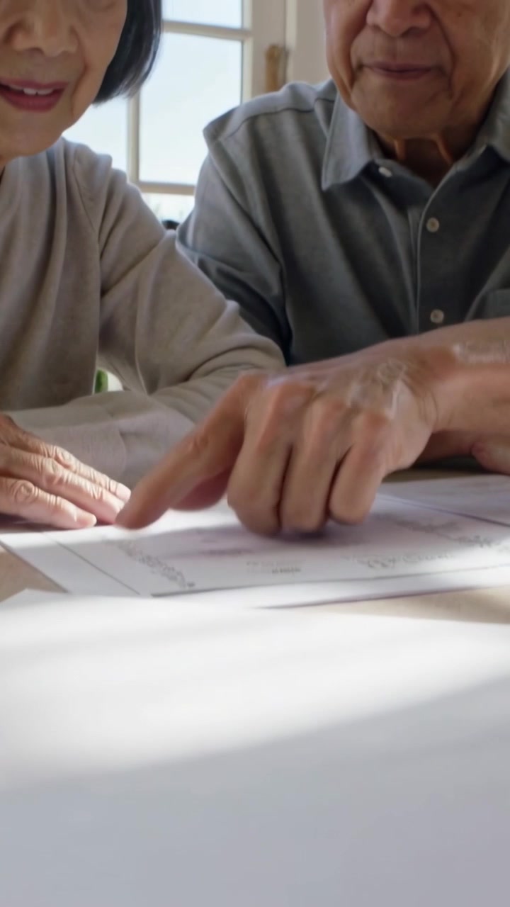 Video verticale: Mano che scorre sui documenti, anziani appoggiati al tavolo che indicano un foglio per la revisione.