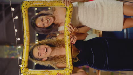 vertical video of female friends having fun posing with photo booth photo frame at party in bar