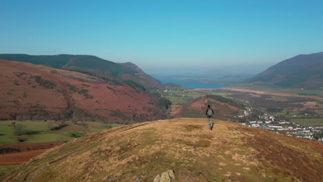 hiker walking to hilltop with slow reveal of green valley and village with mountains in distance at english lake district uk