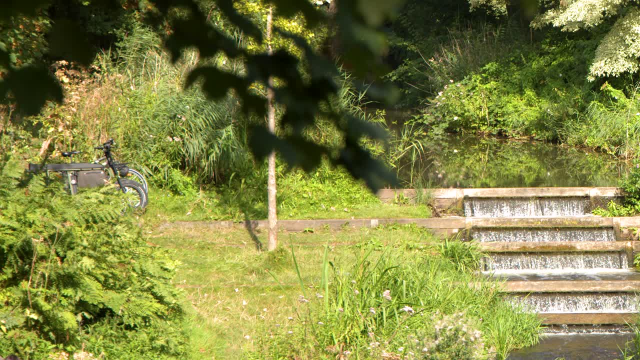 Stationary bicycle near cascading water steps, lush greenery, bright daylight, slight camera movement, tranquil mood