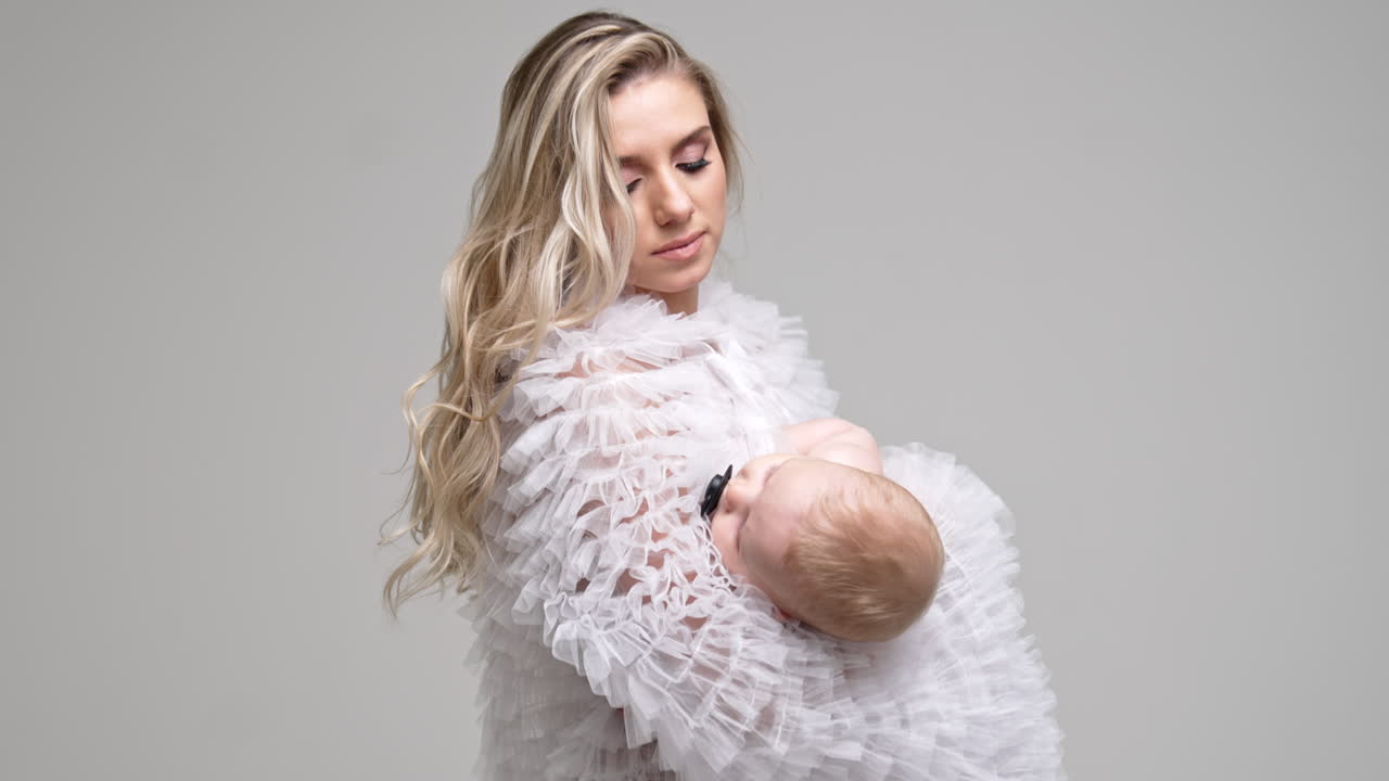 Long-haired woman in white robe holding a baby. Caring mom swaying her child to make him sleep.