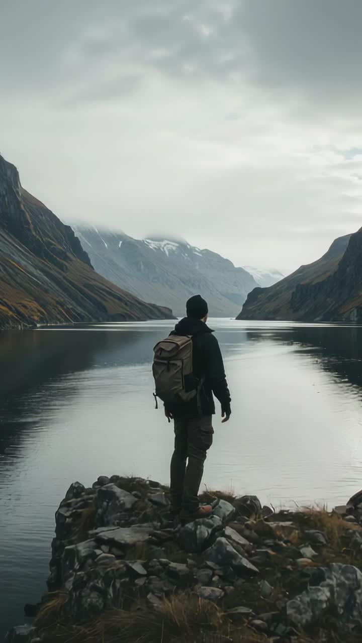 Vertical video: Reaching rock edge, hiker scanning peaks, viewing at mountain lake with pack beanie