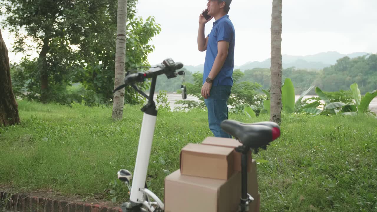 Delivery Asian Man Courier Talking On Mobile Phone With Electric Scooter And Packages For Customers