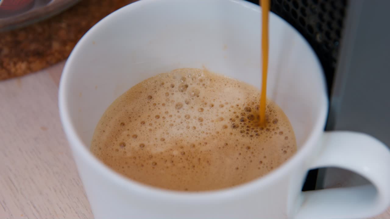 Coffee maker pours hot aroma drink with foam into white ceramic cup standing on wooden table. Domestic kitchen appliance at home close view