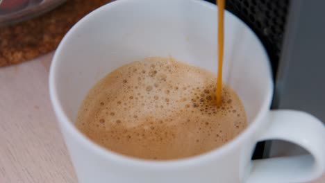 coffee maker pours hot aroma drink with foam into white ceramic cup standing on wooden table. domestic kitchen appliance at home close view