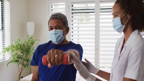 Mixed-race-female-physiotherapist-wearing-mask-helping-senior-exercise-using-dumbbells