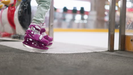 primer plano de una persona entrando en una pista de hielo con patines púrpuras y pantalones de color ceniza, con una vista borrosa de personas patinando en el fondo