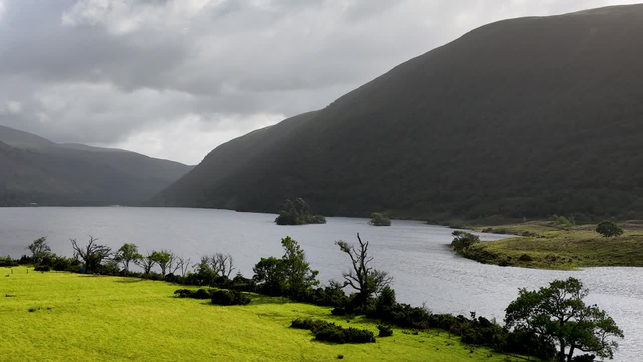 Camera pans across serene lake, green meadow, and mountains under dramatic, overcast natural light