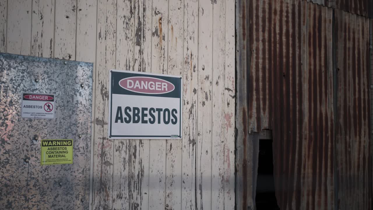 An asbestos hazard warning signs on the weathered exterior of an old industrial building, with barbed wire visible in the foreground