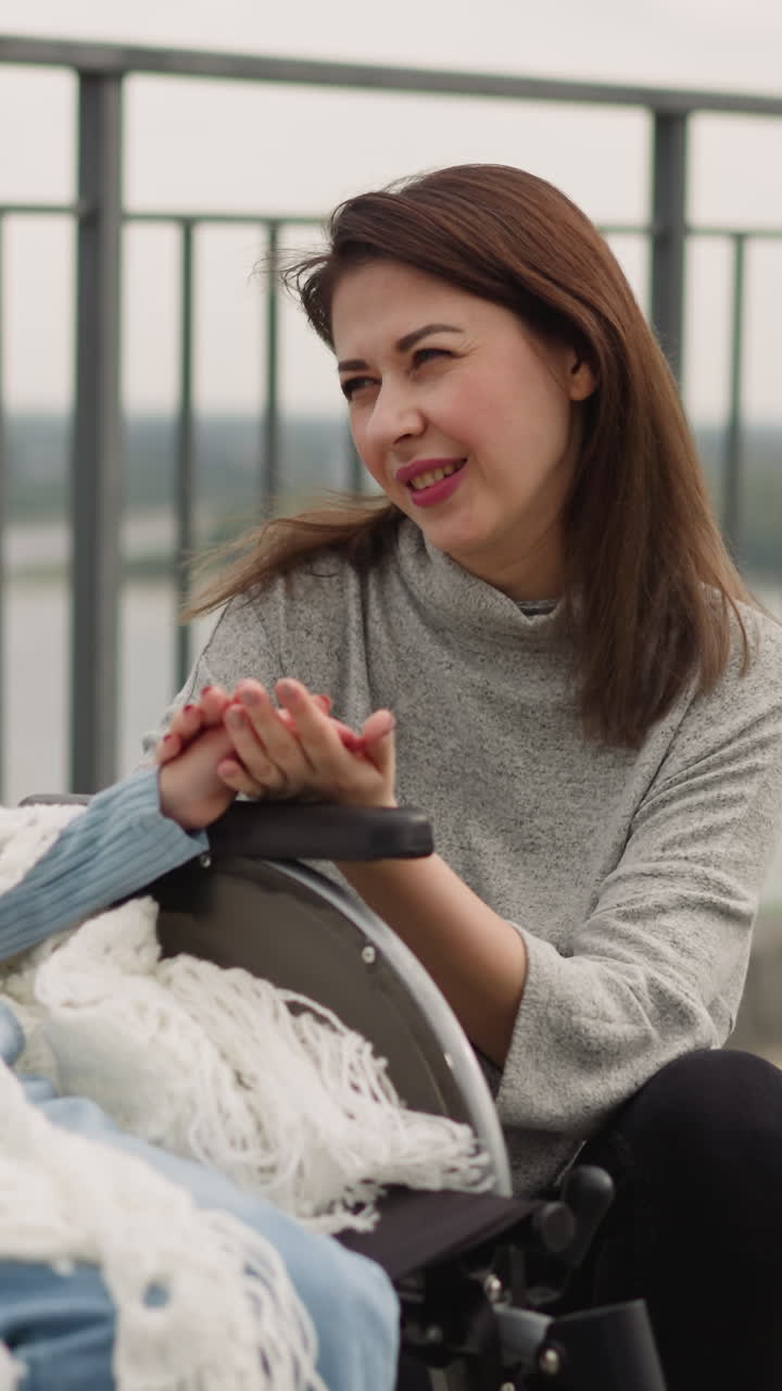 una mujer cuidadosa mira a su hija pequeña con distrofia muscular acariciando suavemente la mano. una niña con un pañuelo se sienta en una silla de ruedas disfrutando de un paseo por la ciudad con su madre