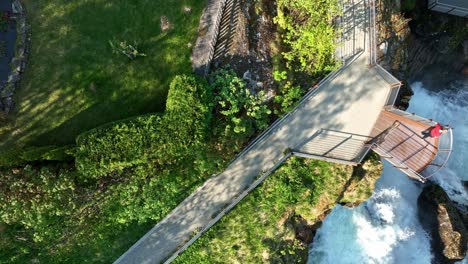 Bird's-Eye-View-of-Majestic-Geiranger-Viewpoint-and-River-Amidst-Norway's-Splendor---Standing-on-platform-above-waterfall-during-sunny-morning-in-Unesco-world-heritage-landscape