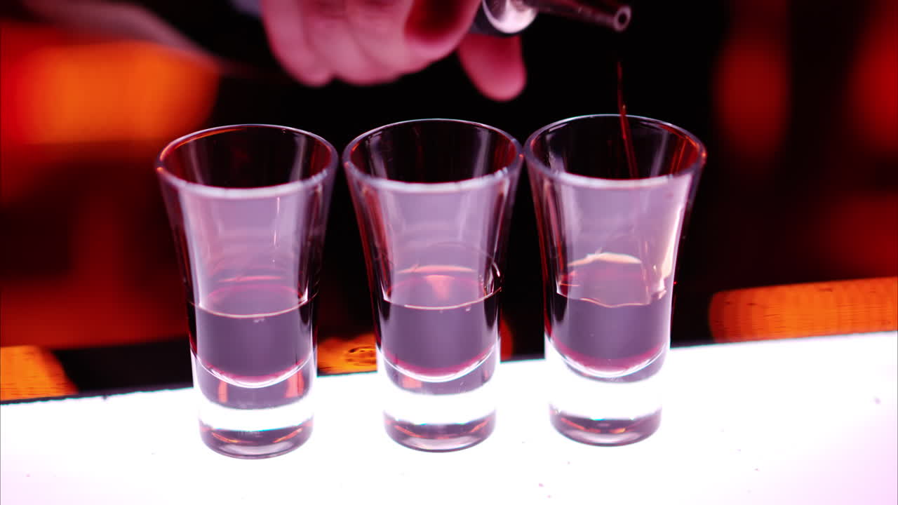Barman making cocktail shots with liquor alcohol in three layers at the bar at night with red neon lights, slow motion