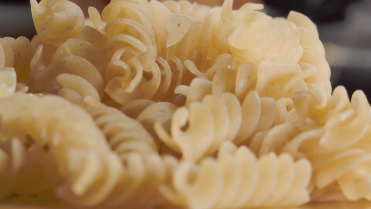 Lähikuva kypsentämättömistä spiraalipastapaloista, jotka esittävät kierteisiä muotoja, uurrettuja pintoja ja vaalean kullanväriä, korostaen yksityiskohtaisesti italialaisen ruoan rakennetta ja kuivapastan ainesosia