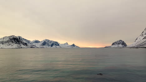flying over fjord water of haukland of lofoten islands in winter season with dramatic sunset in background, aerial drone view