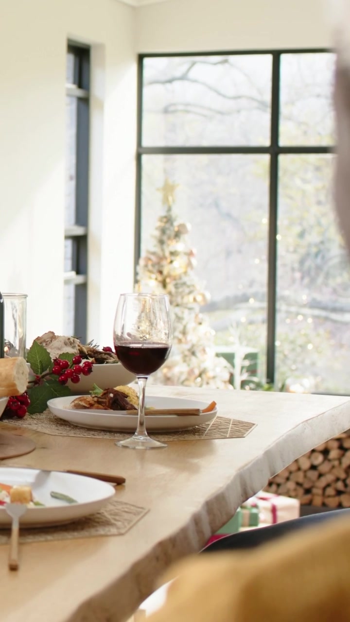 Vertical video: Panning showing man in Santa hat reaching for wine to sip at table, copy space