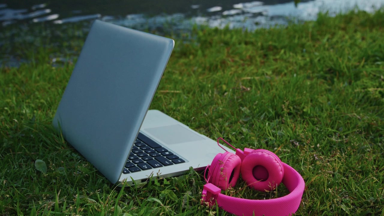 Laptop e cuffie rosa acceso poggiano sull'erba verde vicino all'acqua. Giornata di sole, atmosfera rilassata per lavoro o studio all'aperto. Ideale per contenuti su natura, tecnologia o stile di vita da nomade digitale.