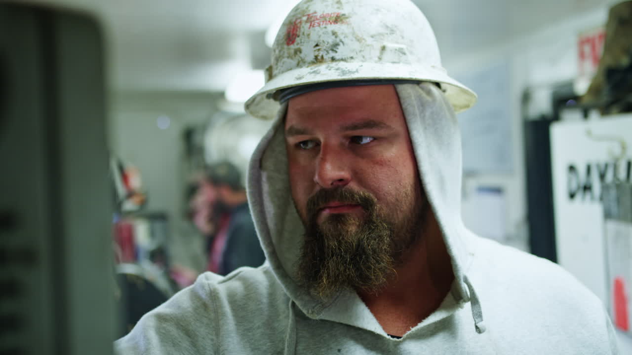 Caucasian man wearing protective helmet and hoodie stands at the wagon with oil drilling equipment. Bearded worker operating the machines controlling oil production.