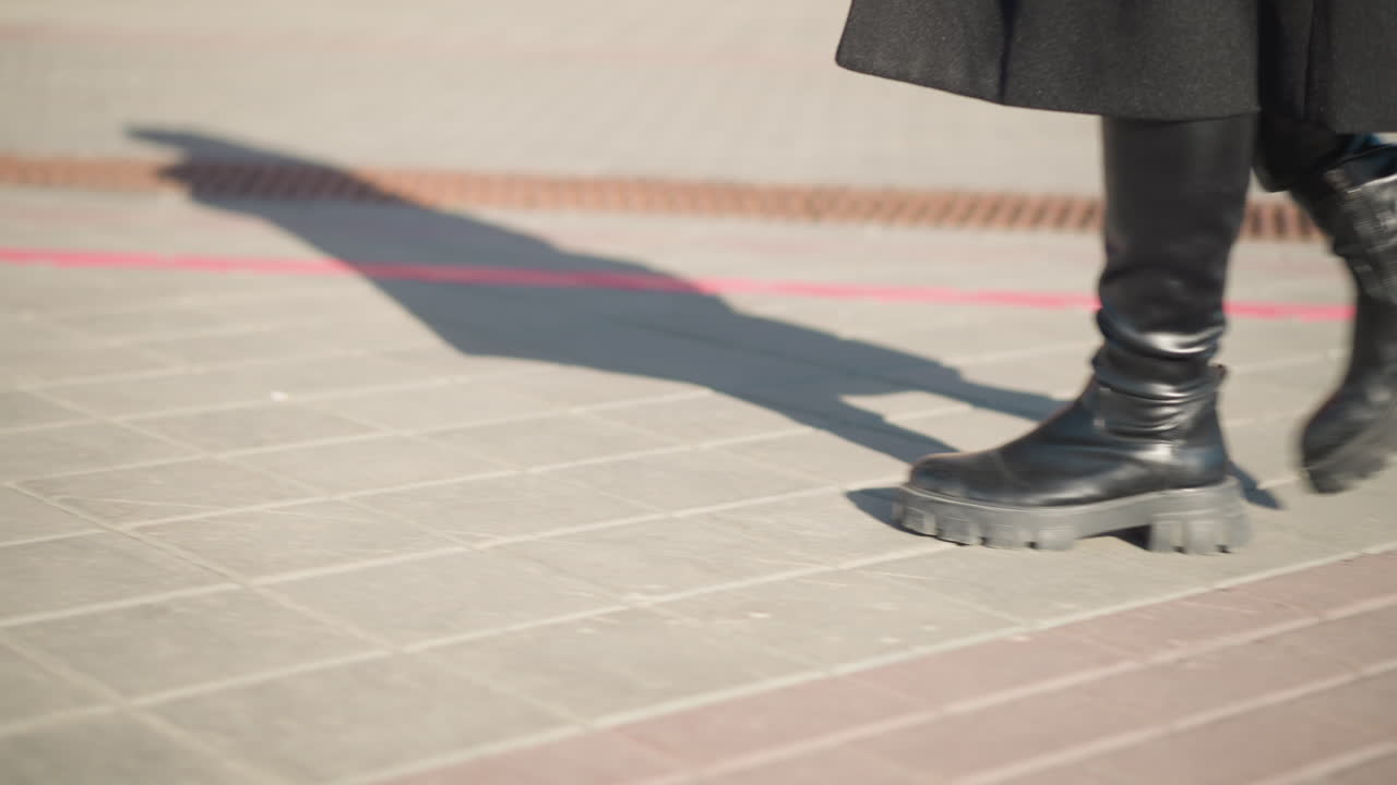 vista de la pierna de un individuo con elegantes botas de cuero y un elegante abrigo negro caminando por el pavimento interconectado iluminado por el sol con largas sombras proyectadas en el suelo