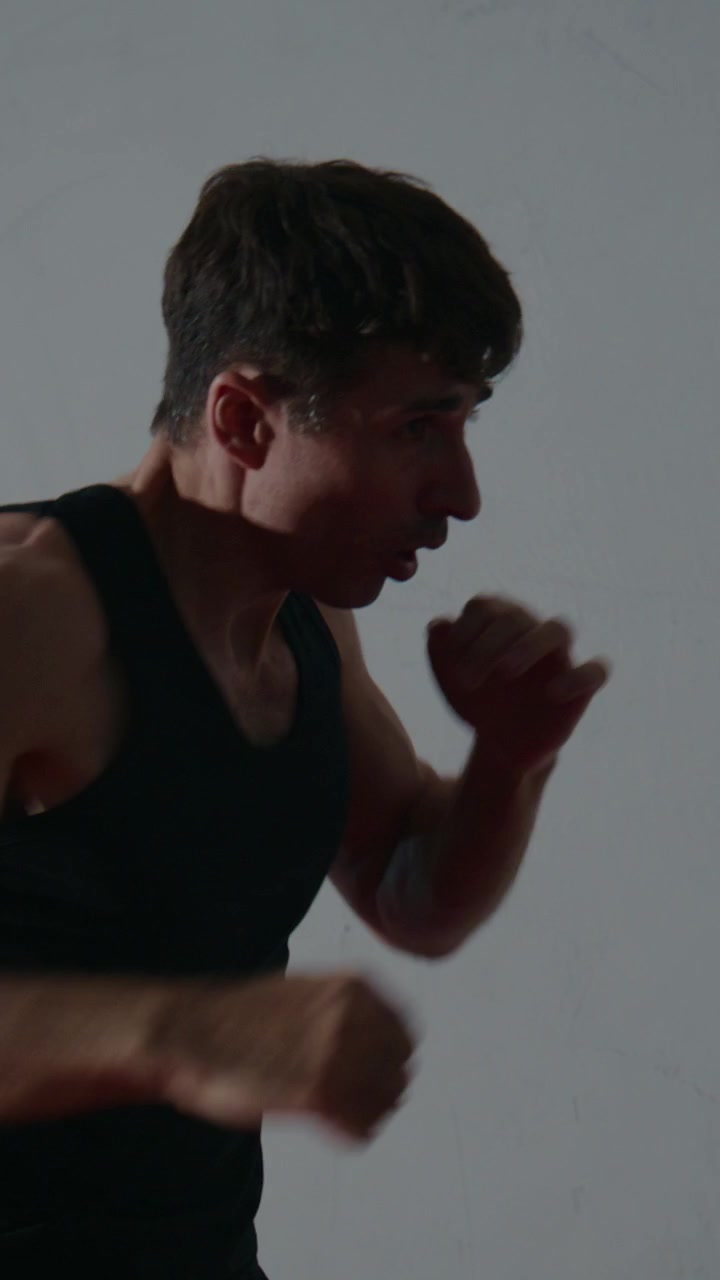 Giovane uomo si allena in posizione da pugilato, concentrato e determinato. La sua ombra si allunga contro una parete semplice, enfatizzando il movimento. Intensa concentrazione visibile nella sua espressione. Perfetto per fitness, sport.