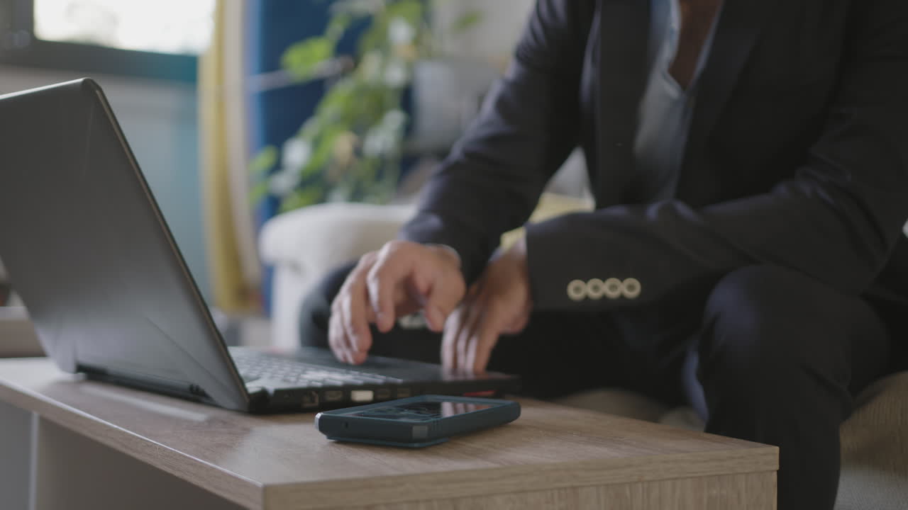 Pop-up do WhatsApp no celular distraindo um jovem empresário trabalhando de casa na sua sala de estar, usando um laptop e vestindo seu terno antes de partir para o voo.