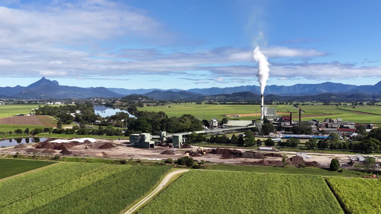 Aerial footage captures a sugar mill in lush fields under clear skies, with smoke rising from the chimney