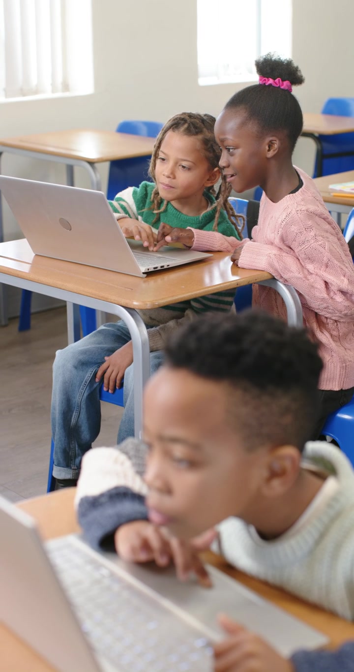Video verticale: Bambini di diverse etnie a scuola seduti ai banchi che condividono i laptop e digitano per un compito.