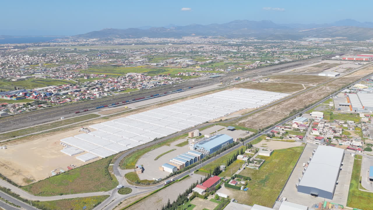 Ampia vista aerea che mostra un grande parco industriale con numerosi magazzini, un hub logistico e un treno merci che passa, sullo sfondo di un vasto paesaggio urbano e montuoso.