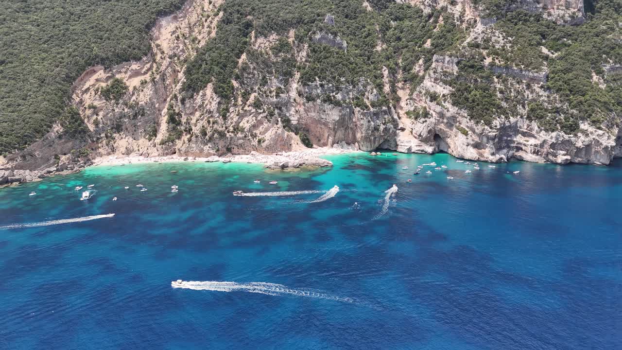 Boats speed across bright blue water near steep cliffs in Sardinia